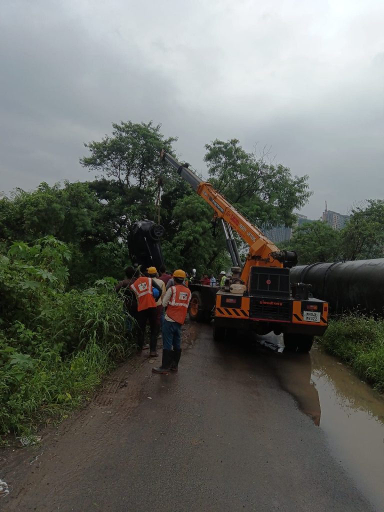 A black overturned car being lifted from a ditch or embankment by a large orange mobile crane, with rescue workers in safety vests on a muddy road.