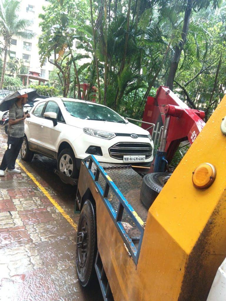 A white Ford EcoSport being lifted by a tow truck on a paved residential lane in Mumbai.