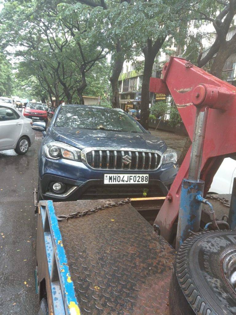 Dark blue Maruti S-Cross being recovered by a tow truck on a wet, tree-lined street in the city during rainfall.