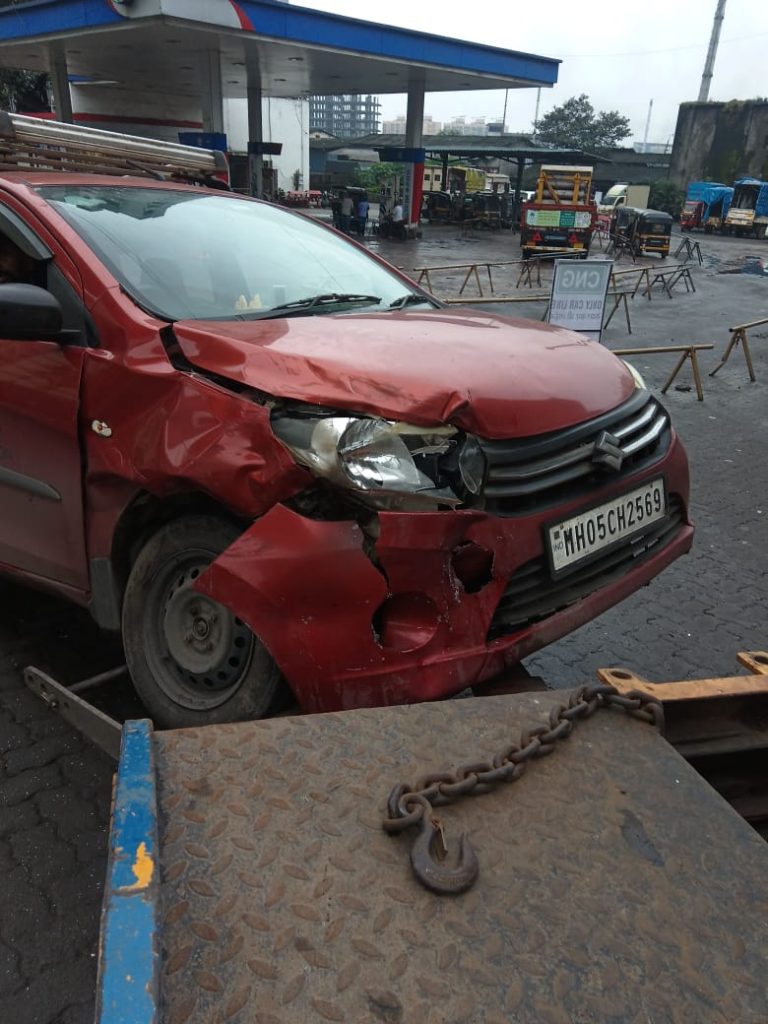 Heavily damaged red Maruti Celerio after an accident, being secured onto a tow truck near a petrol pump.